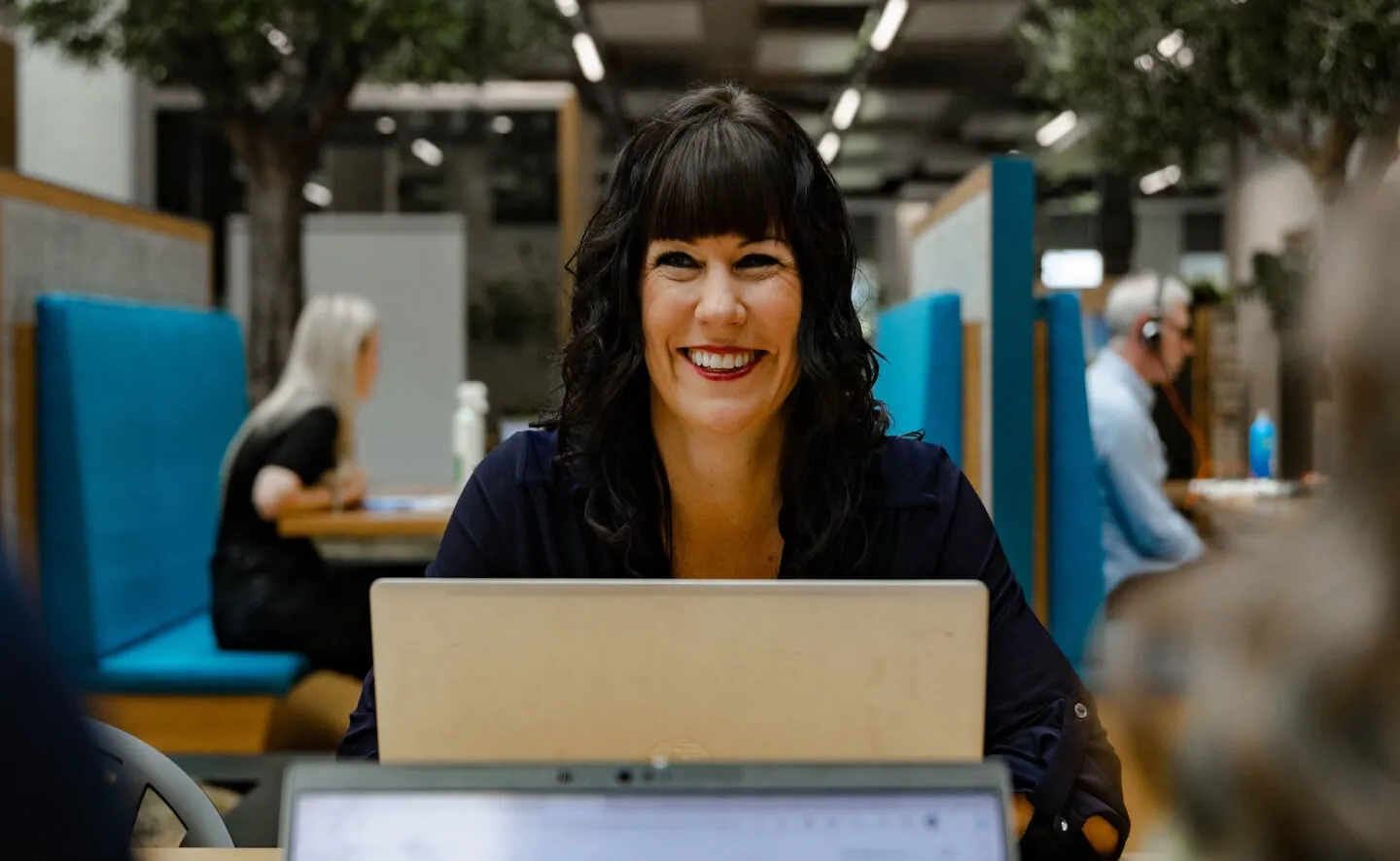 Lady with dark hair smiling as she talks with colleagues whilst working on laptop