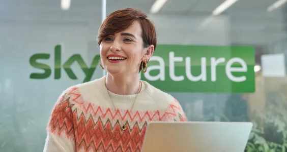 Lady with short red hair talks to a colleague whilst working at her laptop