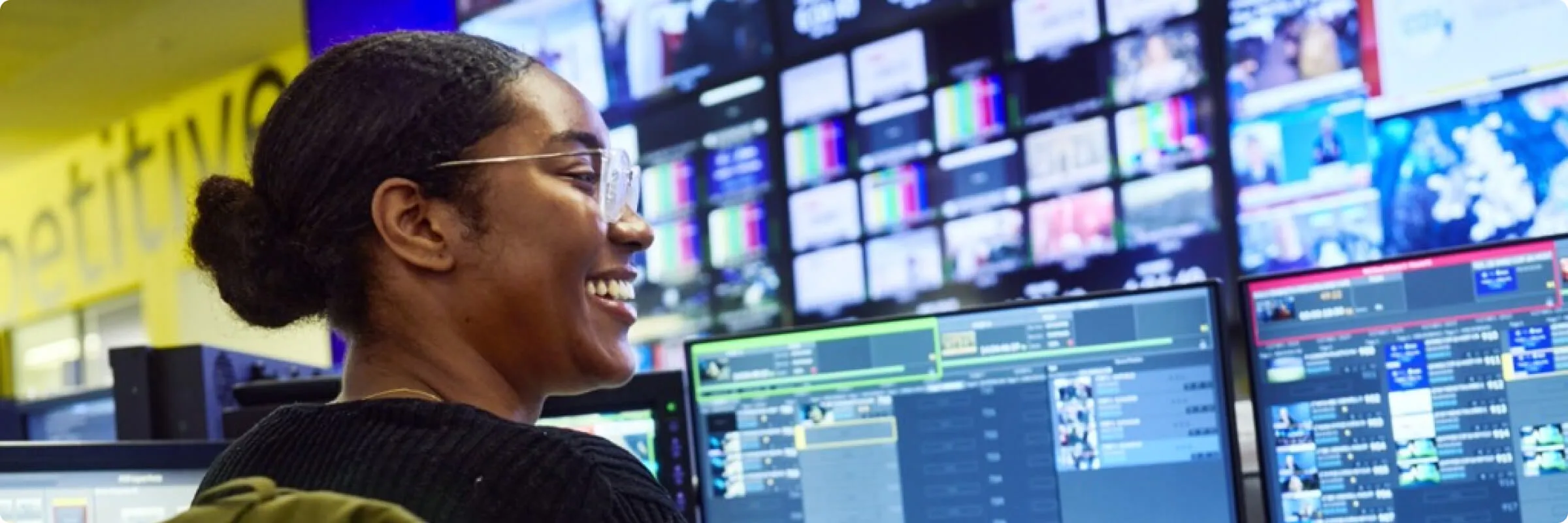 Side view of black lady with glasses and tied up hair smiling whilst working at two screens