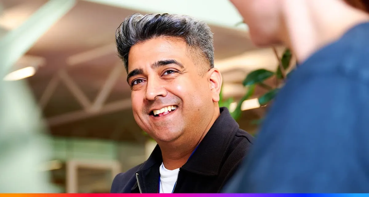 A Smiling Man With Grey Streaked Hair Looks Warmly Toward a Colleague During a Conversation in a Modern Office Environment