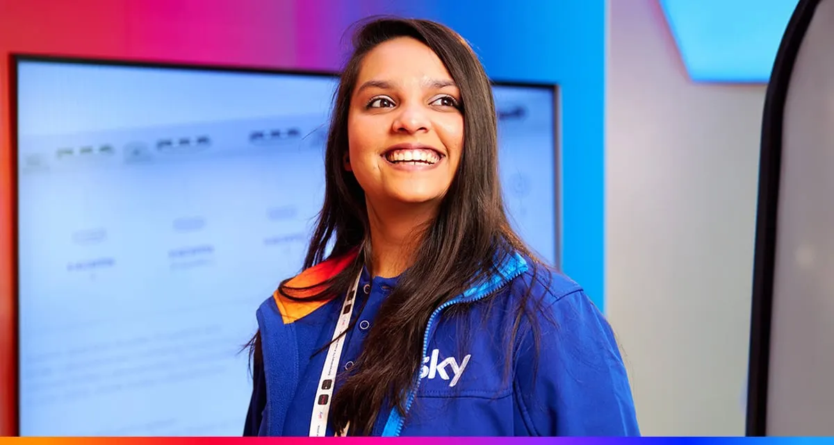 A Smiling Woman Wearing a Blue Sky Branded Jacket Stands in Front of a Brightly Lit Blue and Pink Display Background