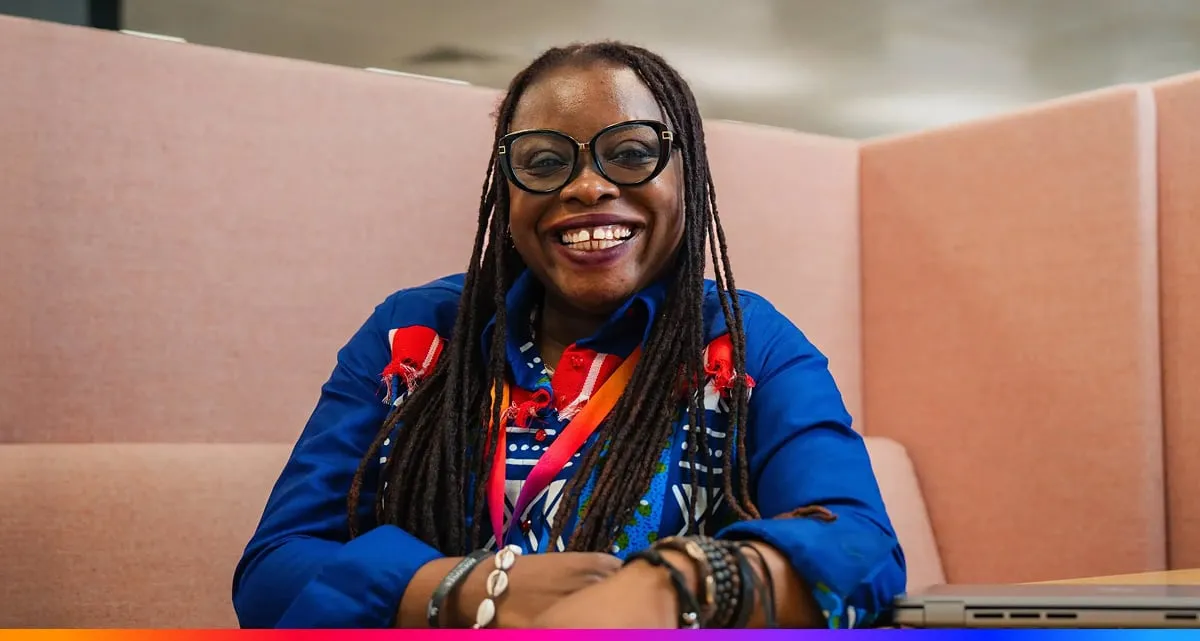 A Smiling Woman With Long Dreadlocks and Glasses Sits in a Pink Acoustic Office Booth Wearing a Vibrant Blue Patterned Shirt